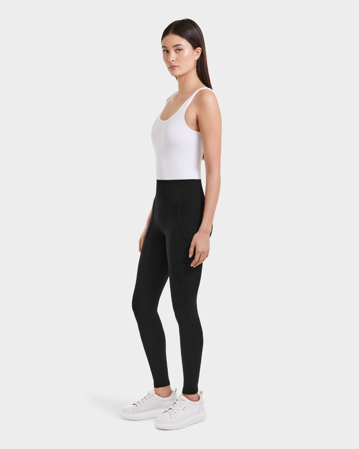A woman stands against a white background, wearing Rudsaks ADELLE - L525010 Black sculpting leggings with pockets, a high-rise waistband, and stretch fabric. She faces slightly to the side with her arms relaxed by her sides.