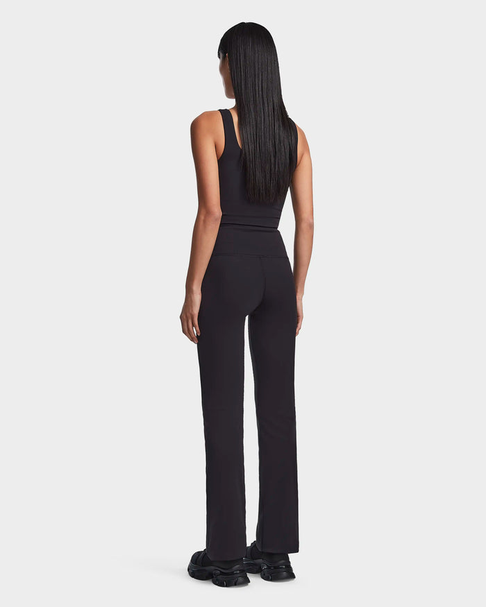 A woman with long straight hair faces away, wearing the Rudsak ADRIANA - 8523550 Black sleeveless top and high-waisted black pants, paired with black sneakers, set against a plain light background.