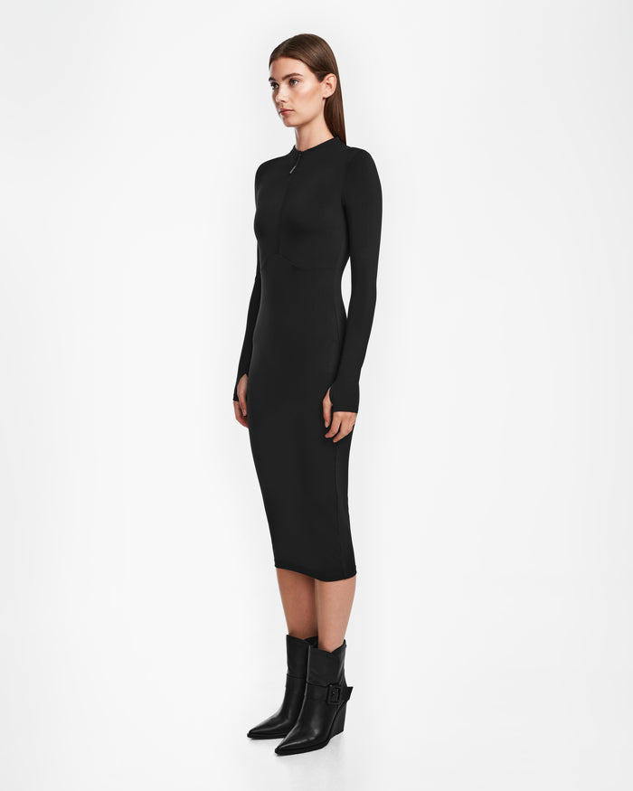 A woman stands in profile against a white background wearing the Rudsak AGNES - 8524551 Black fitted, long-sleeve zip-up front dress paired with black ankle boots featuring a small heel.