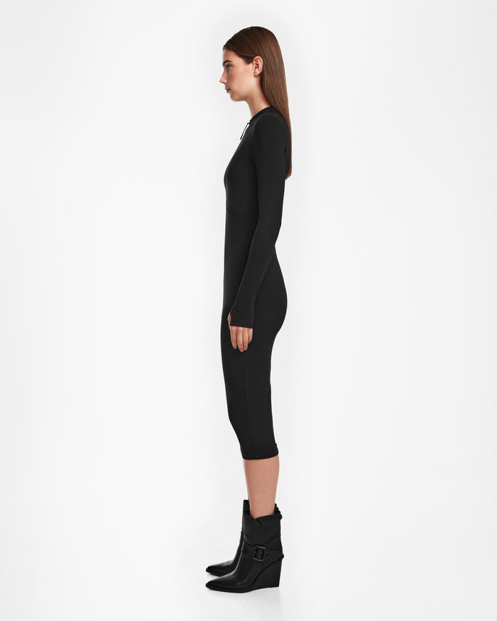 A woman with straight brown hair stands in profile wearing the Rudsak AGNES - 8524551 Black, a fitted, long-sleeve women’s bodycon stretch dress, paired with black buckled ankle boots, against a plain white background.