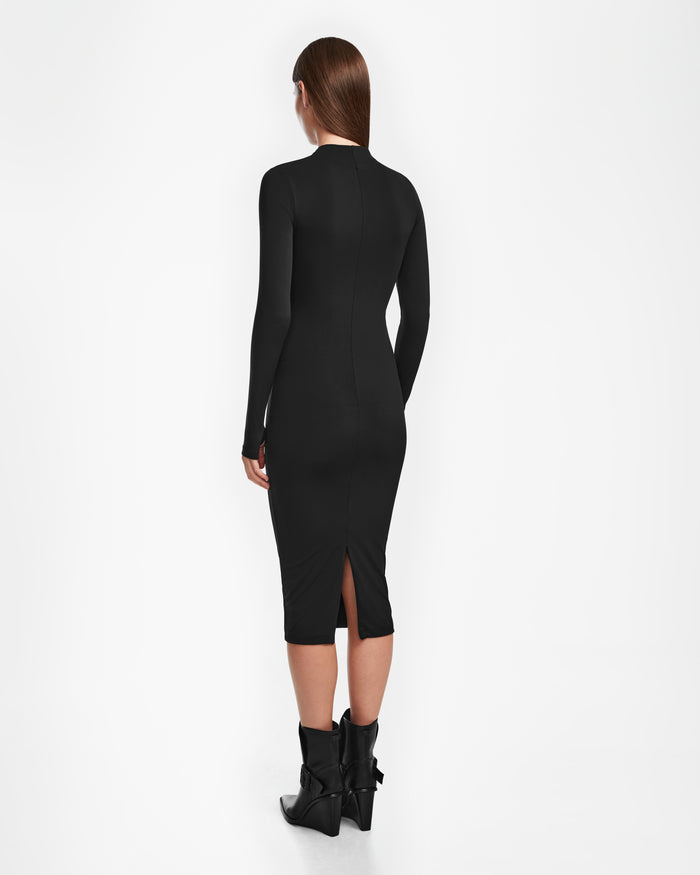 A woman with straight brown hair models the Rudsak AGNES - 8524551 Black, a fitted, long-sleeve black bodycon stretch dress with a back slit. She stands with her back to the camera and pairs it with black ankle boots featuring bows.