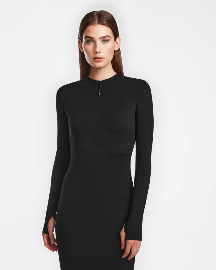 A woman with straight brown hair wears the Rudsak AGNES - 8524551 Black dress, a fitted, long-sleeved style with a zip-up front. She stands against a plain white background, looking directly at the camera with a neutral expression.