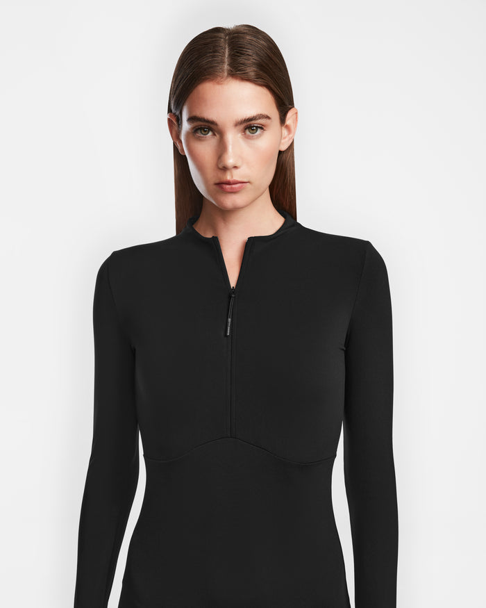 A woman with straight brown hair wears the Rudsak AGNES - 8524551 Black fitted zip-up dress with long sleeves, standing against a white background and looking directly at the camera.