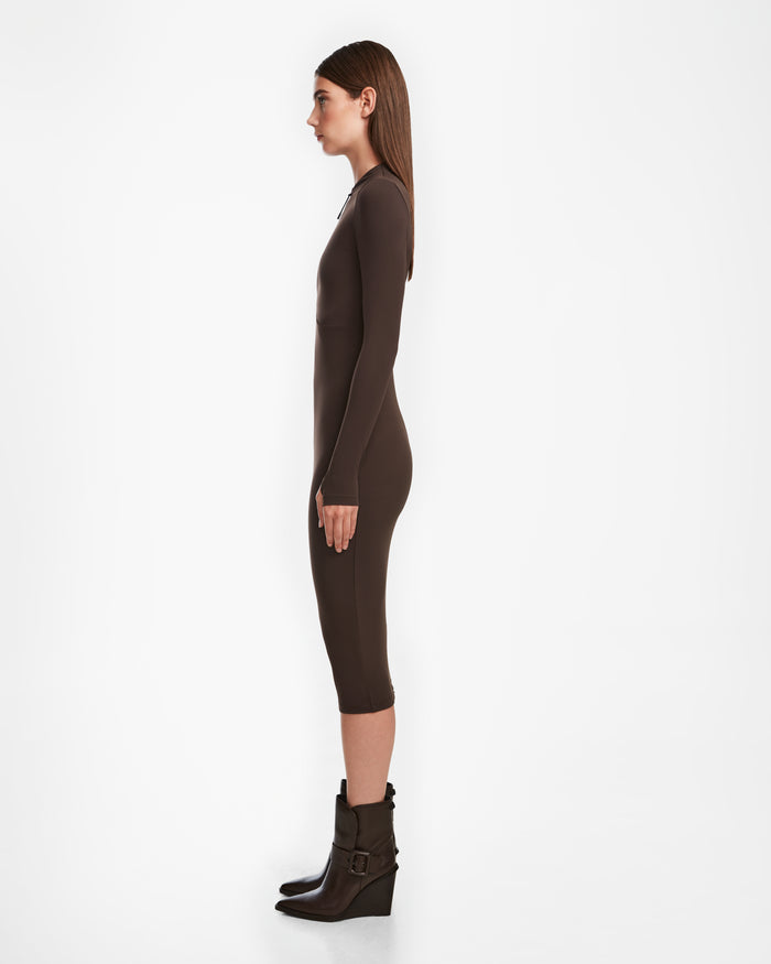 A woman in profile against a white background wears a fitted, long-sleeve bodycon dress that falls below the knees and pairs it with Rudsaks AGNES - 8524551 Espresso ankle boots.