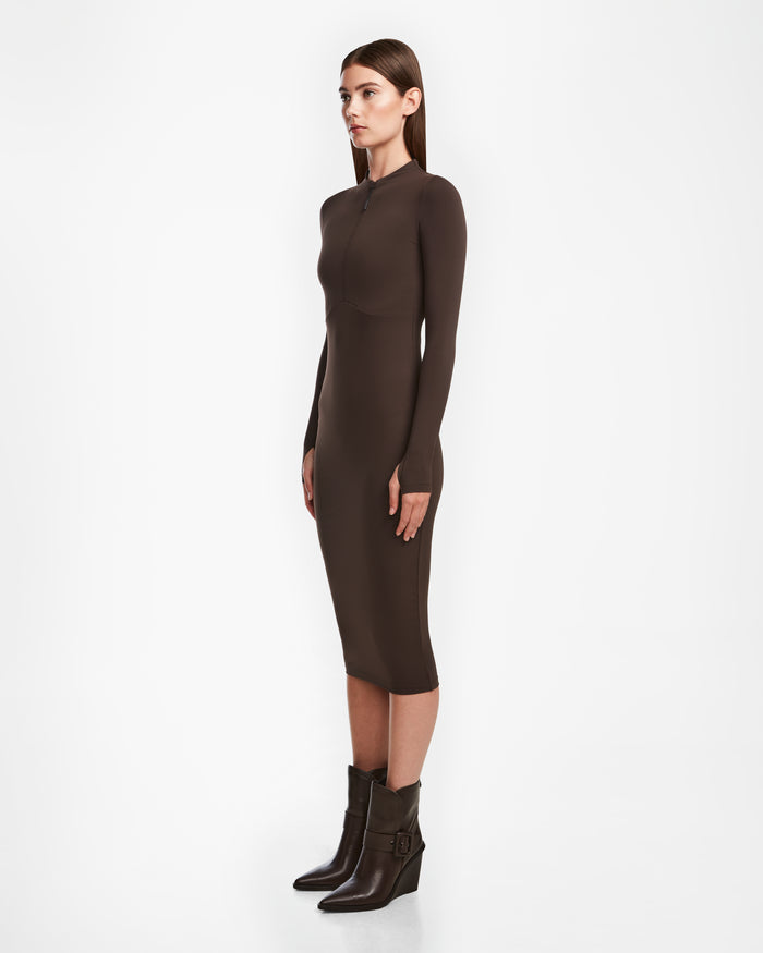 A woman stands in profile against a plain white background wearing the fitted, long-sleeve AGNES - 8524551 Espresso bodycon dress by Rudsak, paired with matching brown ankle boots featuring buckles.