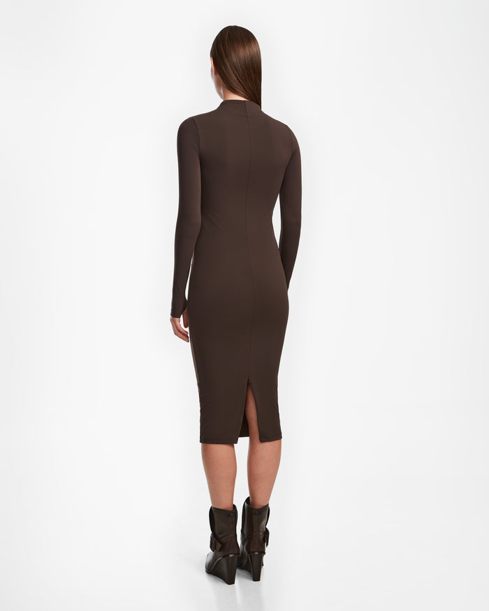 A woman with straight brown hair stands with her back to the camera in a fitted Rudsak AGNES - 8524551 Espresso bodycon dress with long sleeves and a rear slit, styled with black ankle boots against a clean white background.