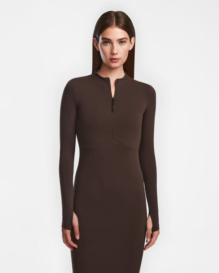 A woman with straight brown hair wears the Rudsak AGNES - 8524551 zip-up fitted dress in Espresso, standing against a plain white background.
