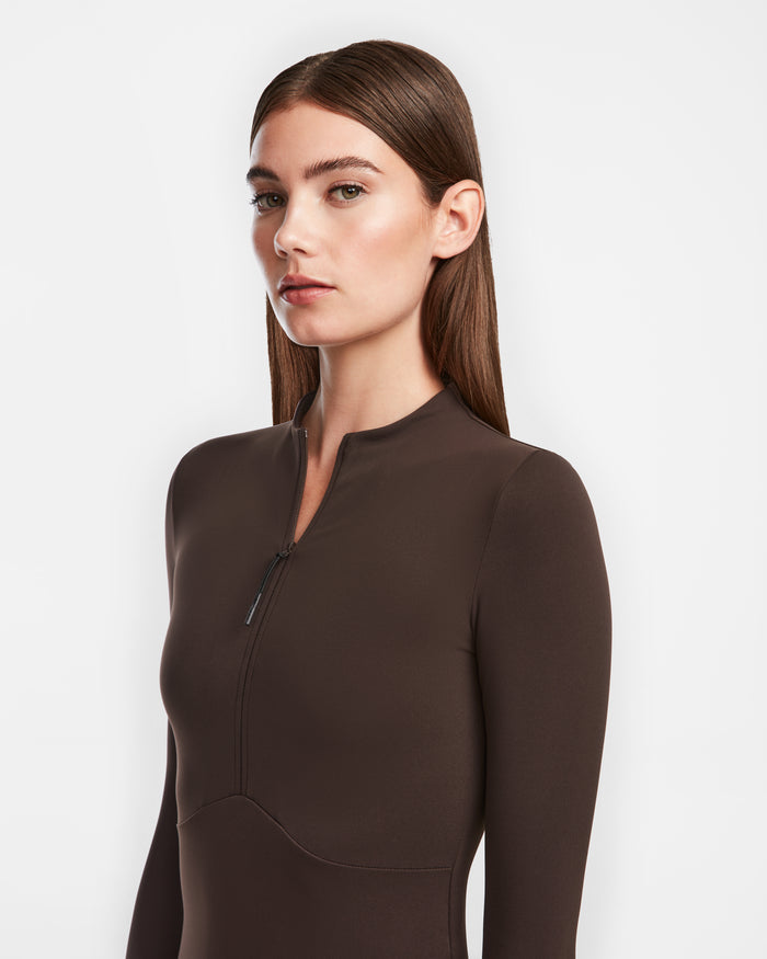 A woman with straight brown hair models the Rudsak AGNES - 8524551 Espresso, a dark brown zip-up long-sleeve fitted stretch dress, standing against a plain white background and looking slightly left.