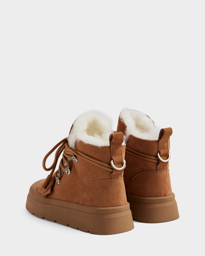 The Rudsak ALEXEY - L225025 Tan is a women’s suede winter boot in brown, featuring plush wool lining, a thick rubber sole, metal eyelets, and back pull tabs, shown from a rear side angle.