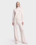 A person stands facing forward against a plain white background, wearing the ALISYA - 8526505 Iceland set by Rudsak: a cozy cream-colored high-neck sweater featuring a small “R” on the chest, paired with matching wide-leg pants.