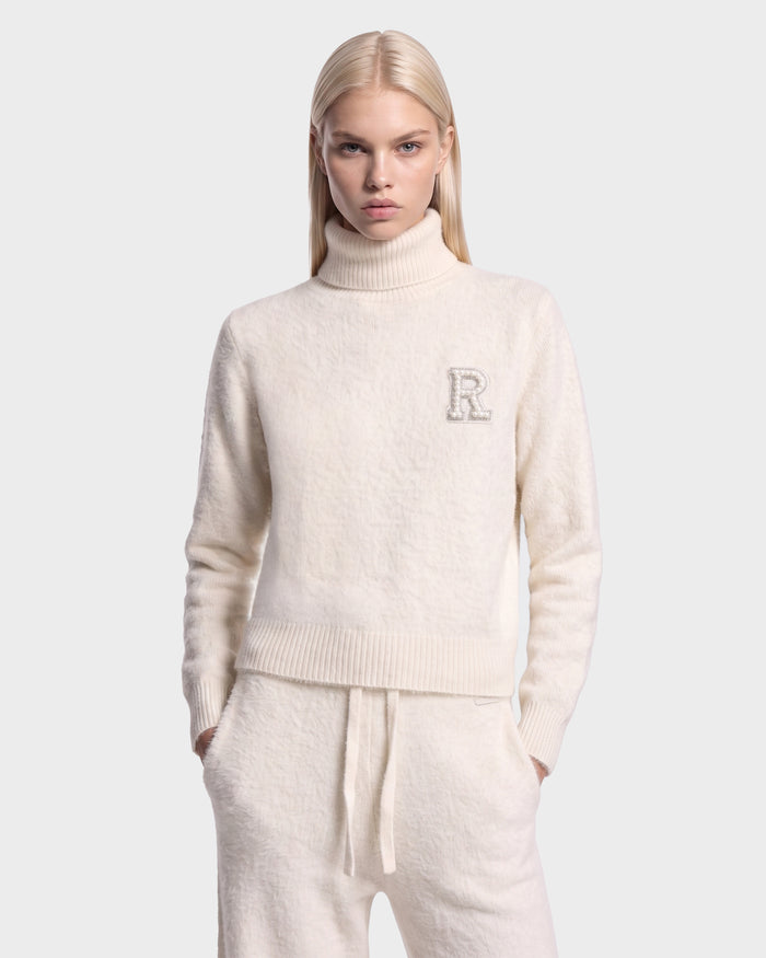A person with long blonde hair wears the ALISYA - 8526505 Iceland cream-colored turtleneck sweater with an R patch on the left chest and matching drawstring pants by Rudsak, standing against a plain light gray background.