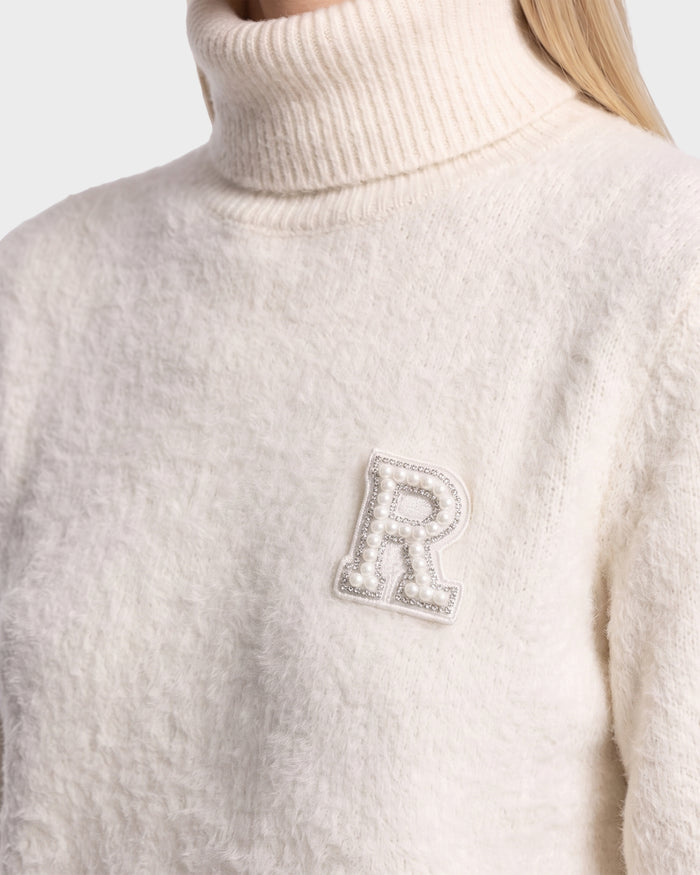 A person wearing the ALISYA - 8526505 Iceland textured white turtleneck sweater by Rudsak, featuring a decorative letter R patch adorned with pearls on the left side of the chest.