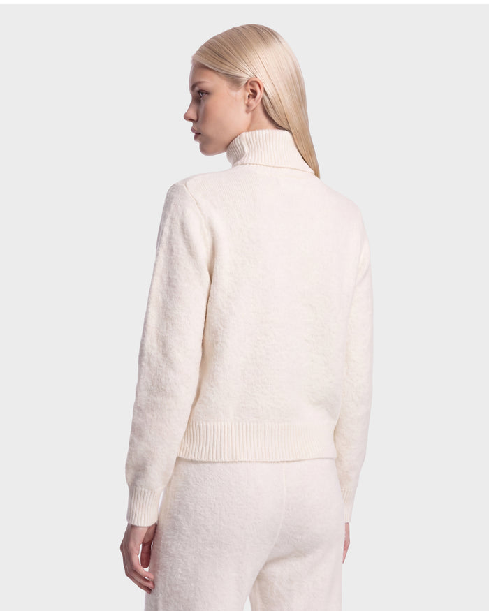 A person with straight, light blond hair wears the Rudsak ALISYA - 8526505 Iceland cream-colored turtleneck sweater and matching pants, standing against a plain light gray background, facing away and looking slightly to the side.