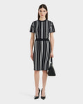 A woman in the Rudsak ANGELA - 8525006 Black dress with white vertical stripes stands against a white background, styled with black heels and a coordinating handbag. Her straight dark hair and neutral expression enhance her modern, elegant look.