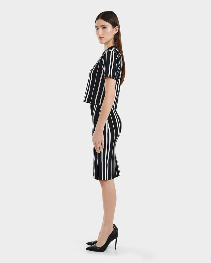 A woman stands in profile against a white background, wearing the Rudsak ANGELA - 8525006 Black dress—short-sleeved, knee-length with white vertical stripes—and black heels, embodying modern elegance and a flattering silhouette.