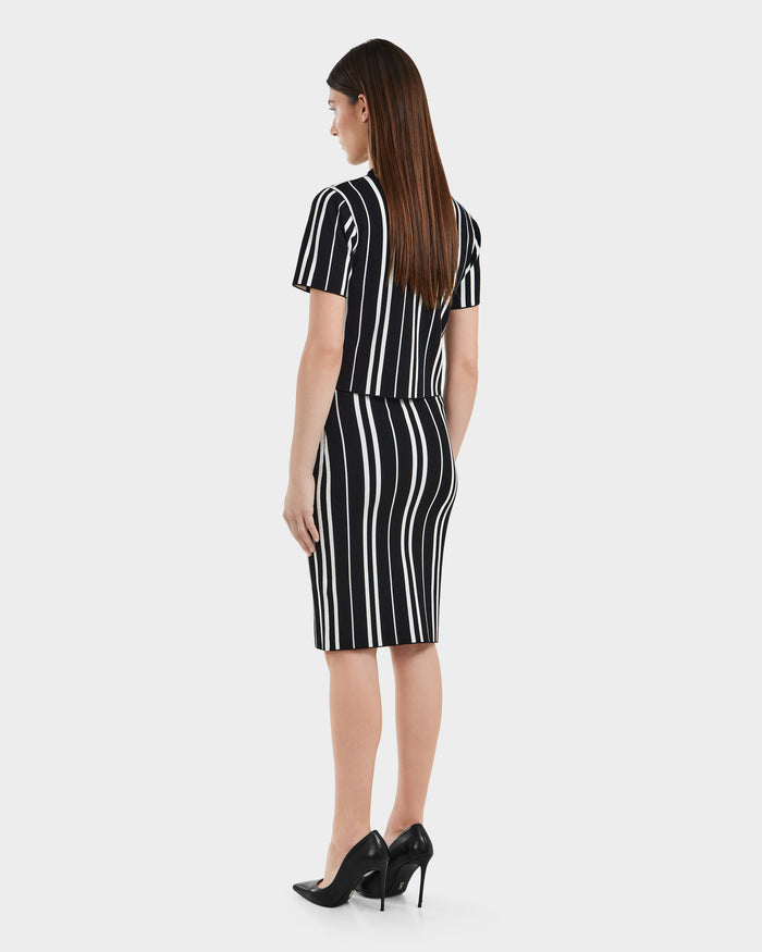 A woman stands facing away in the Rudsak ANGELA - 8525006 Black dress, featuring white vertical stripes, short sleeves, and a knee-length hem; she pairs it with black heels against a plain white background.