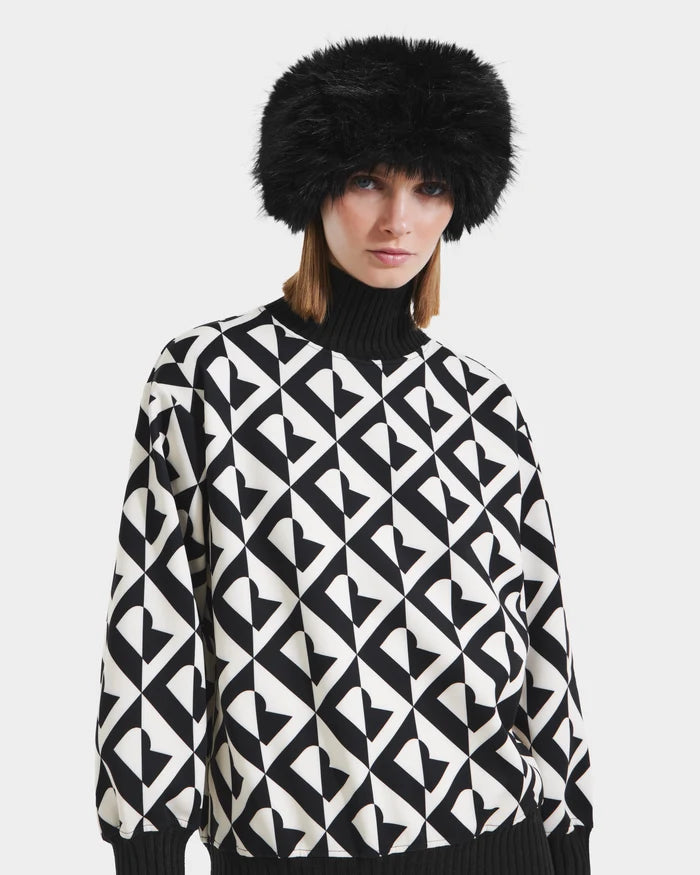 A woman models the Rudsak ARLETTE - 8425544 Black faux fur hat, paired with a black-and-white geometric heart-patterned sweater, creating a cozy-chic look against a plain white background.