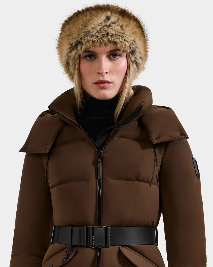 Wearing the Rudsak ARLETTE - 8425544 Mix Mocha puffer coat with a black belt and faux fur-trimmed hood, this look captures cozy-chic winter style against a simple, light backdrop.