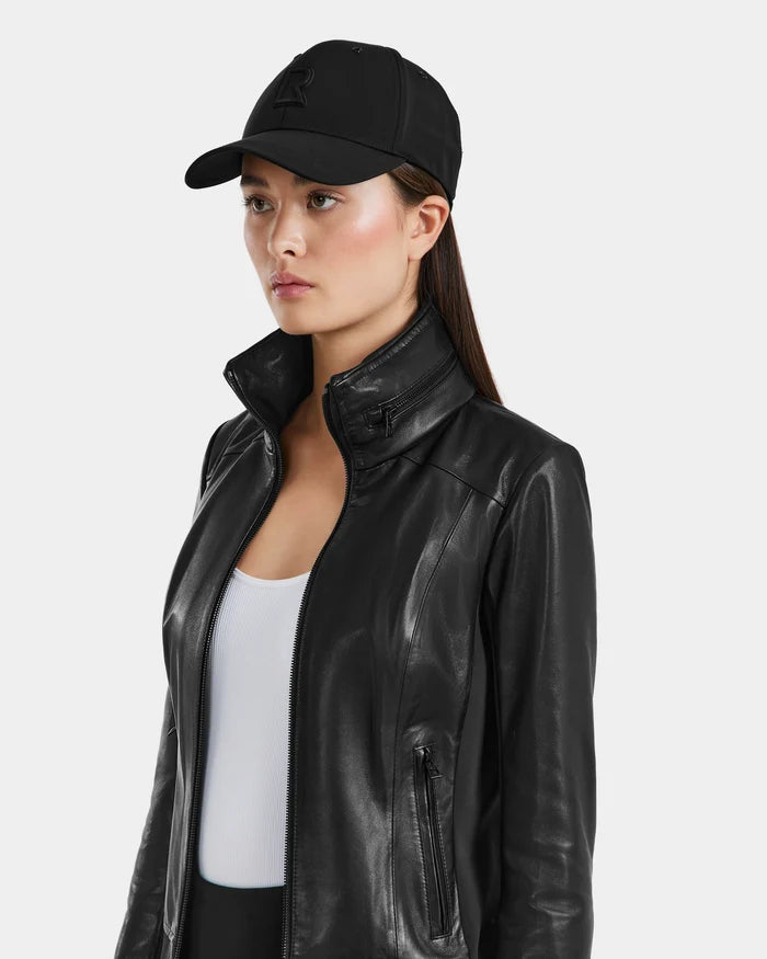 A woman wearing a black Rudsak ARSENE - 9422504 cap, black leather jacket, and white top stands against a plain white background, gazing to the side with a neutral expression.
