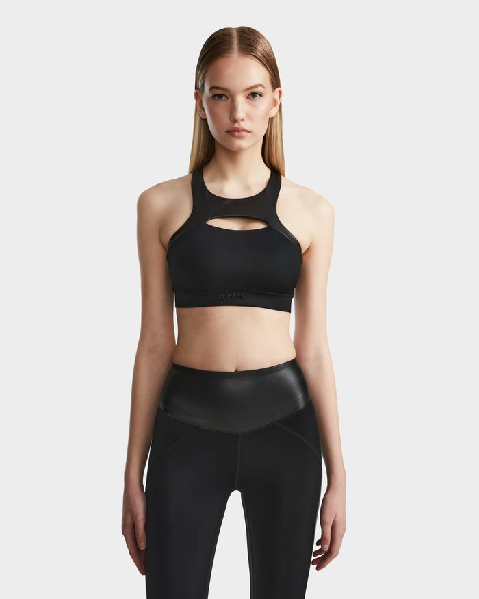 A woman with straight, light brown hair wears the RUDSAK ARYA - 8523002 BLACK sports bra and high-waisted black leggings, standing against a plain light gray background.