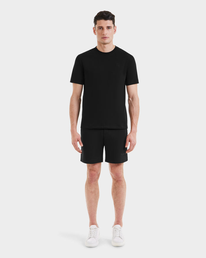 A man faces forward wearing the Rudsak ASHER - M525000 Black tailored-fit mens cotton t-shirt with a minimalist chest logo, paired with black shorts and white sneakers, set against a plain white background.
