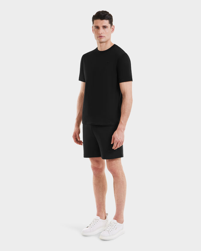 A man stands against a plain white background, wearing the Rudsak ASHER - M525000 Black mens cotton t-shirt with a minimalist chest logo, paired with black shorts and white sneakers. He faces slightly to the side with a neutral expression.