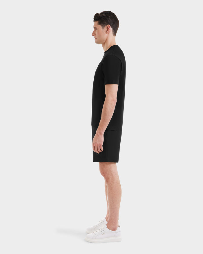 A man in profile against a white background wears the Rudsak ASHER - M525000 Black tailored fit men’s cotton t-shirt with a minimalist chest logo, paired with black shorts and white sneakers.