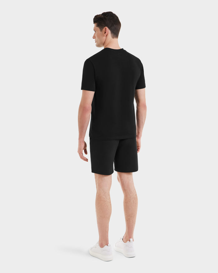A man faces away, wearing the Rudsak ASHER - M525000 Black mens tailored-fit cotton t-shirt with a minimalist chest logo, paired with black shorts and white sneakers against a plain white background.