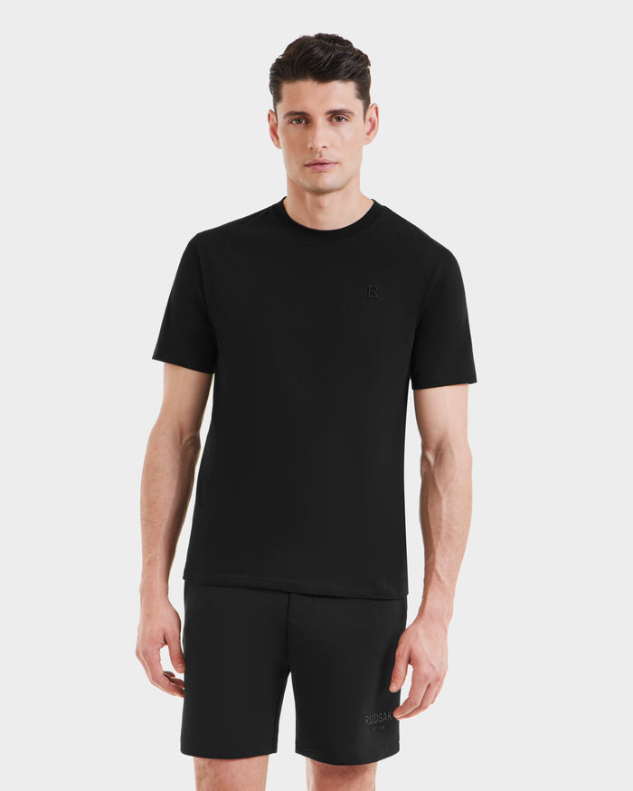 A man with short brown hair stands against a plain white background, looking straight ahead in a neutral expression, wearing Rudsak’s ASHER - M525000 Black men’s cotton t-shirt and black shorts.