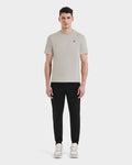 A man faces forward against a plain white background, wearing Rudsaks ASHER - M525000 Cream tailored-fit mens cotton T-shirt with a minimalist chest logo, paired with black jogger pants and beige sneakers.