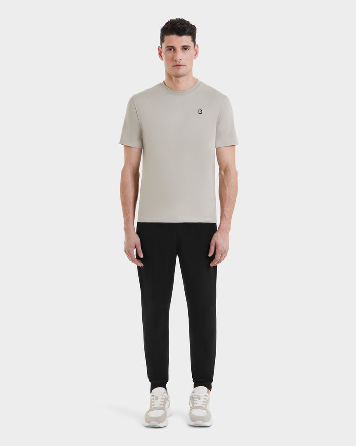 A man faces forward against a plain white background, wearing Rudsaks ASHER - M525000 Cream tailored-fit mens cotton T-shirt with a minimalist chest logo, paired with black jogger pants and beige sneakers.