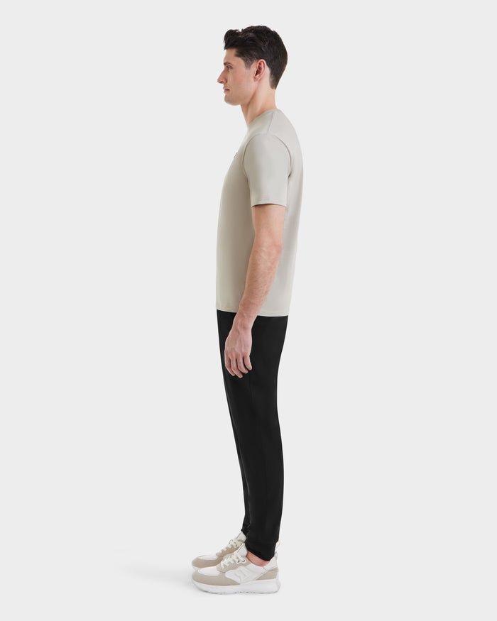 A man in profile faces left, wearing the Rudsak ASHER - M525000 Cream tailored fit men’s cotton T-shirt with a minimalist chest logo, black pants, and beige sneakers, set against a plain white background.
