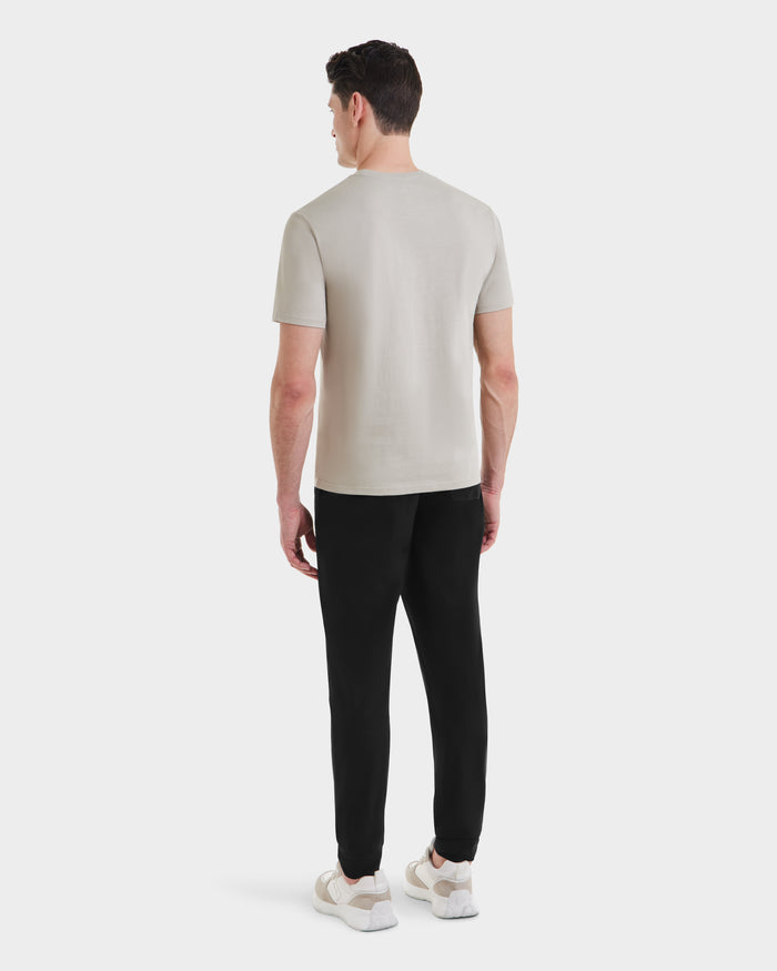 A man seen from behind wears the Rudsak ASHER - M525000 Cream mens cotton t-shirt with a tailored fit, paired with black pants and white sneakers, standing against a plain white background.