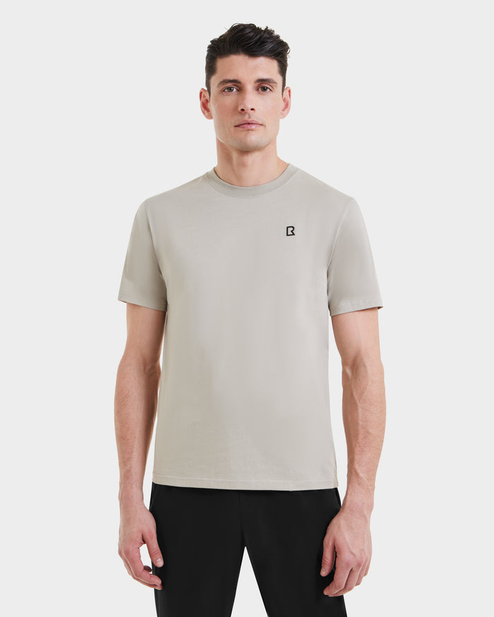A man stands against a plain white background wearing the Rudsak ASHER - M525000 Cream tailored fit men’s cotton t-shirt with a minimalist chest logo, paired with black pants.