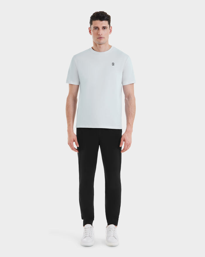 A man faces forward wearing the Rudsak ASHER - M525000 Sky Blue men’s cotton t-shirt with a minimalist chest logo, paired with fitted black pants and white sneakers, all set against a plain white background.