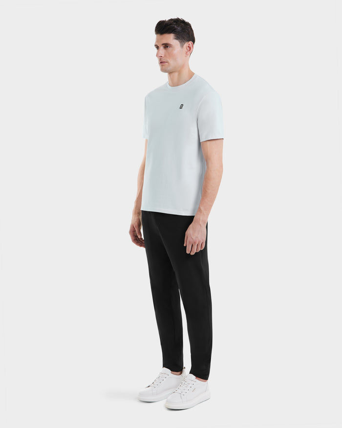 A man faces slightly left, wearing the Rudsak ASHER - M525000 Sky Blue tailored fit mens cotton t-shirt with a minimalist chest logo, paired with black pants and white sneakers, against a white background.