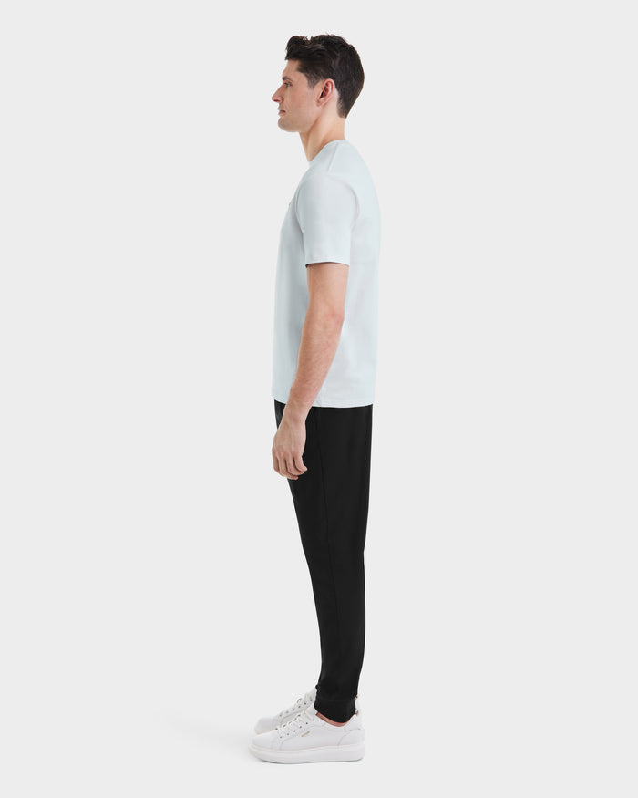 A man in profile facing left wears the Rudsak ASHER - M525000 Sky Blue tailored mens cotton T-shirt with a minimalist chest logo, paired with black pants and white sneakers, set against a plain white background.