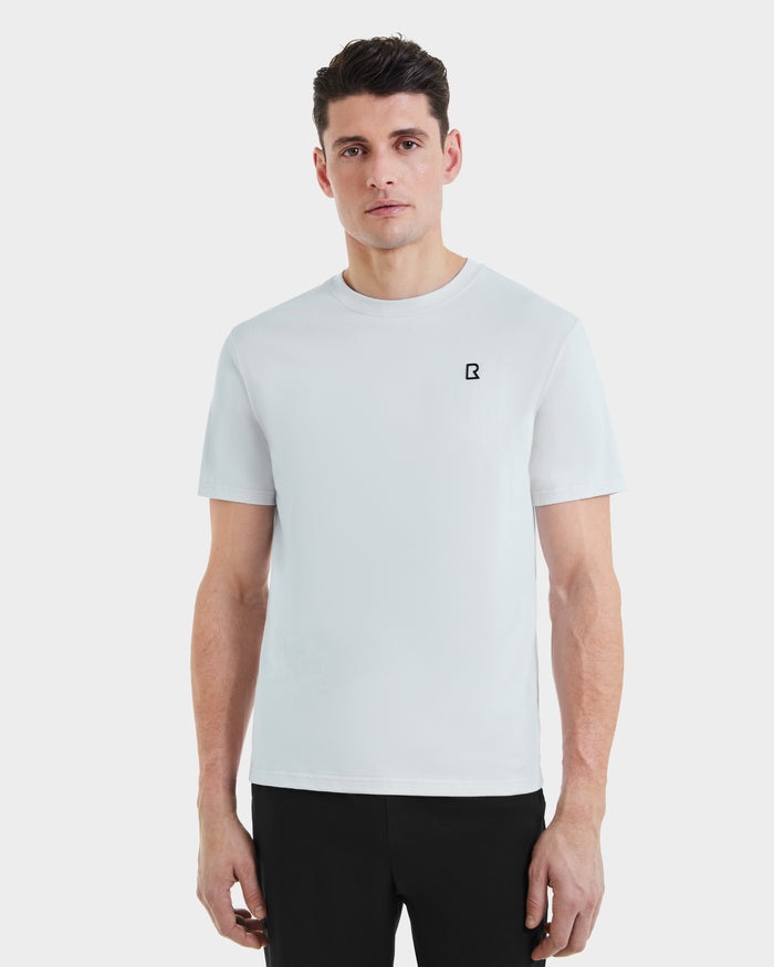 A man with short dark hair wears the Rudsak ASHER - M525000 Sky Blue mens cotton t-shirt with a minimalist chest logo and tailored fit, paired with black pants, standing against a white background.