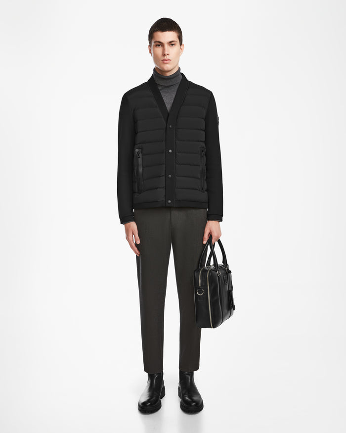 A man stands against a white background wearing the Rudsak ATOM - 6124591 Black hybrid jacket over a gray turtleneck, dark trousers, and black shoes, holding a black leather bag in his left hand.