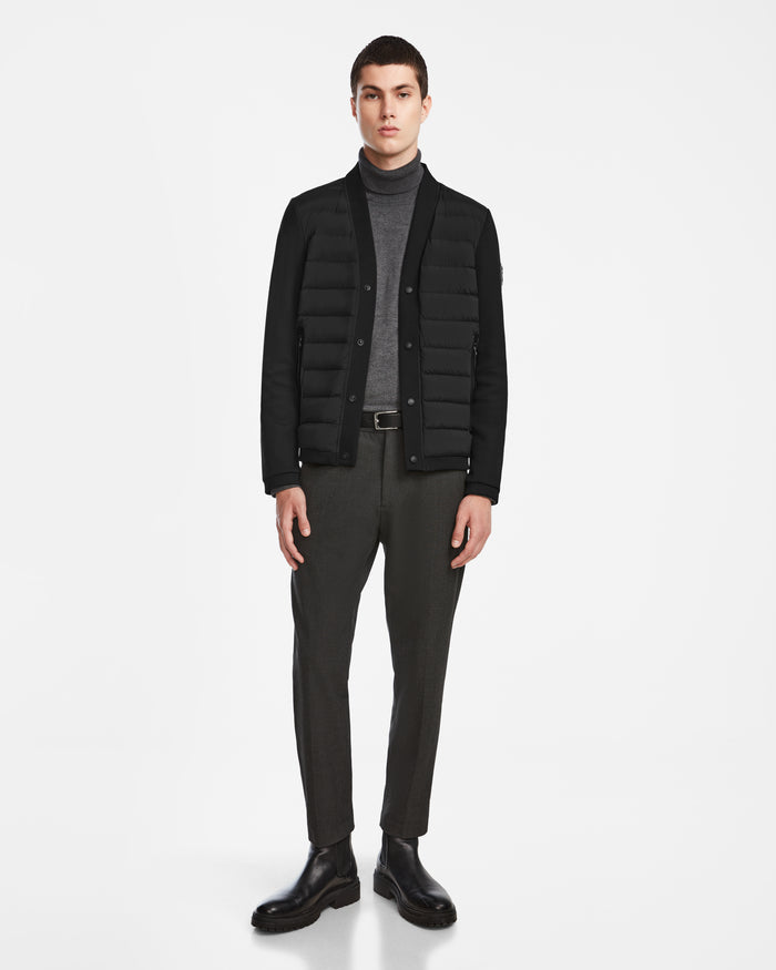 A man stands against a white background wearing the Rudsak ATOM - 6124591 Black hybrid jacket over a gray turtleneck, dark trousers, and black boots. He faces forward with a neutral expression and relaxed posture.