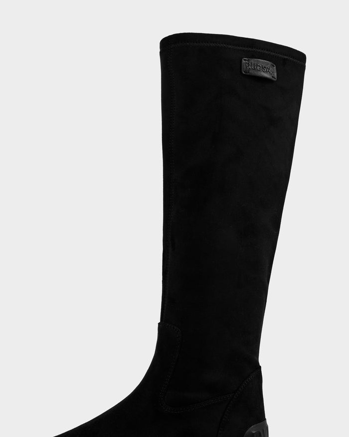 A close-up of the AVOLA - L225514 Black women’s knee-high faux-suede boot by Rudsak, featuring a subtle logo patch near the top, photographed against a plain light gray background.