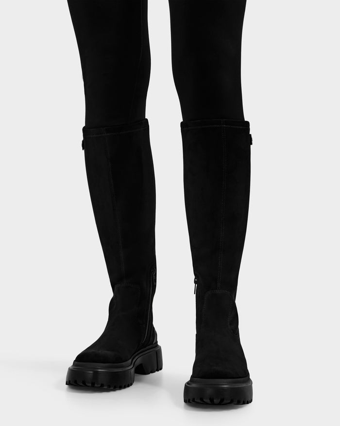 A person wearing the Rudsak AVOLA - L225514 Black women’s knee-high boots with chunky soles, paired with black fitted pants, standing on a plain white background.