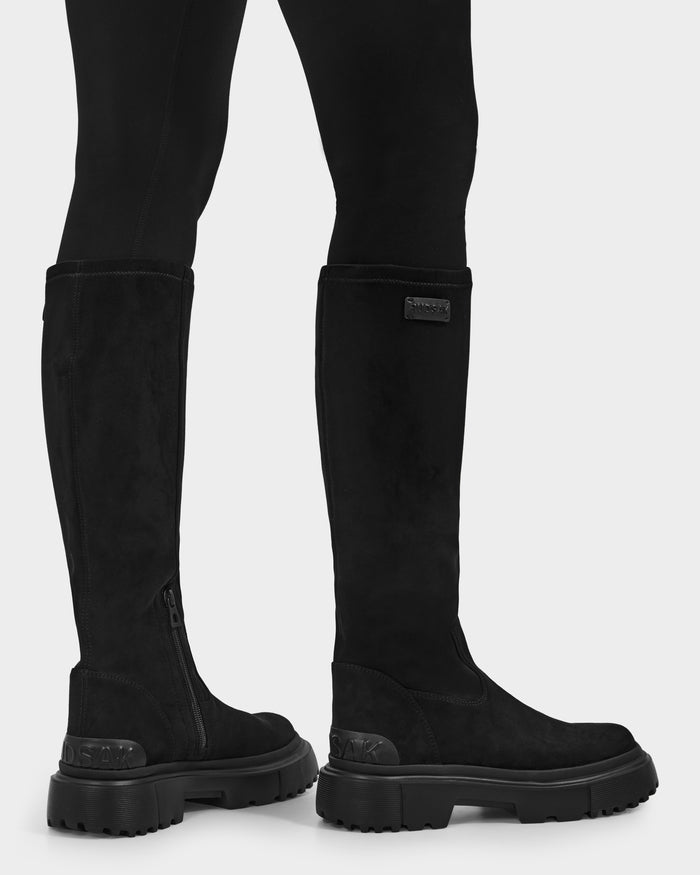 A person stands on a light background wearing Rudsak’s AVOLA - L225514 Black women’s knee-high boots with thick soles and side zippers, paired with black leggings.