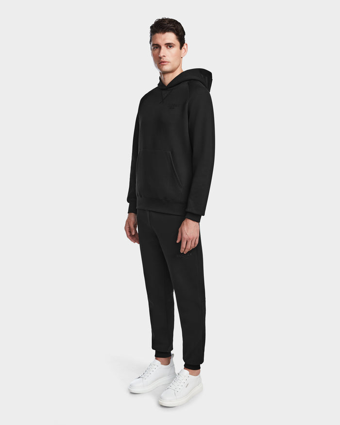 Someone stands against a plain light background wearing Rudsaks BARRY - 9524011 Black M oversized unisex hoodie, paired with matching black joggers and white sneakers.