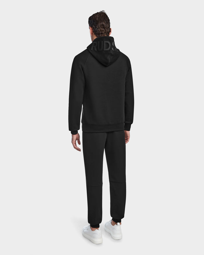 A man stands with his back to the camera in a black Rudsak BARRY - 9524011 hoodie, hood up, and black jogger pants. The unisex fit is paired with white sneakers, set against a plain light background.