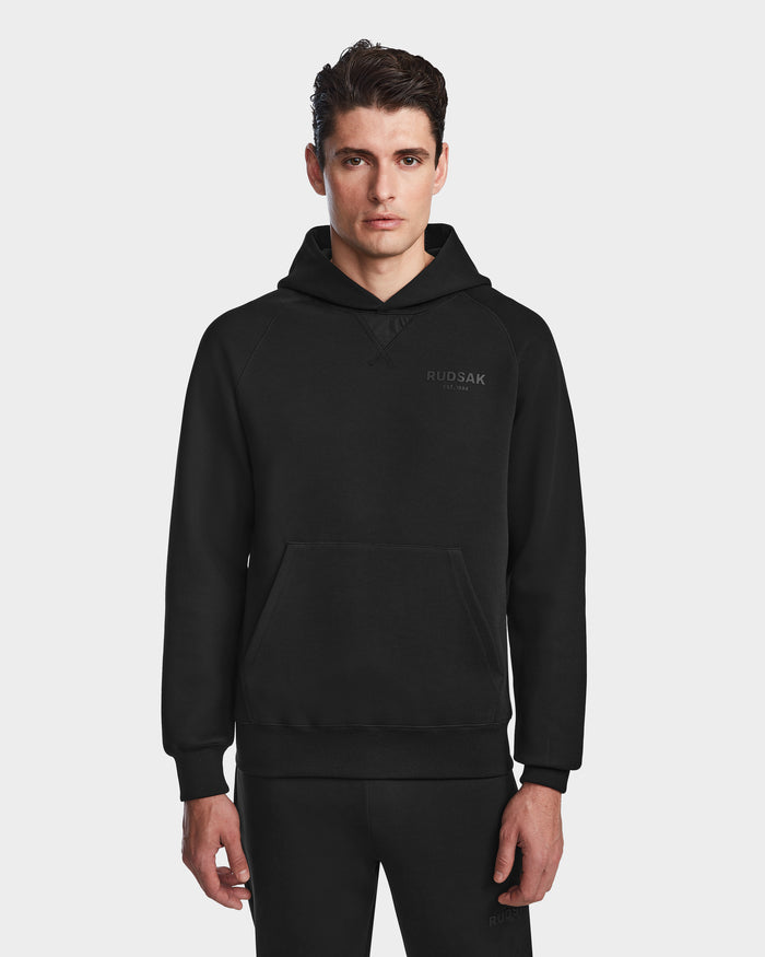 A young man with short dark hair wears the Rudsak BARRY - 9524011 Black M hoodie and matching pants, standing against a plain white background, facing forward with a neutral expression.
