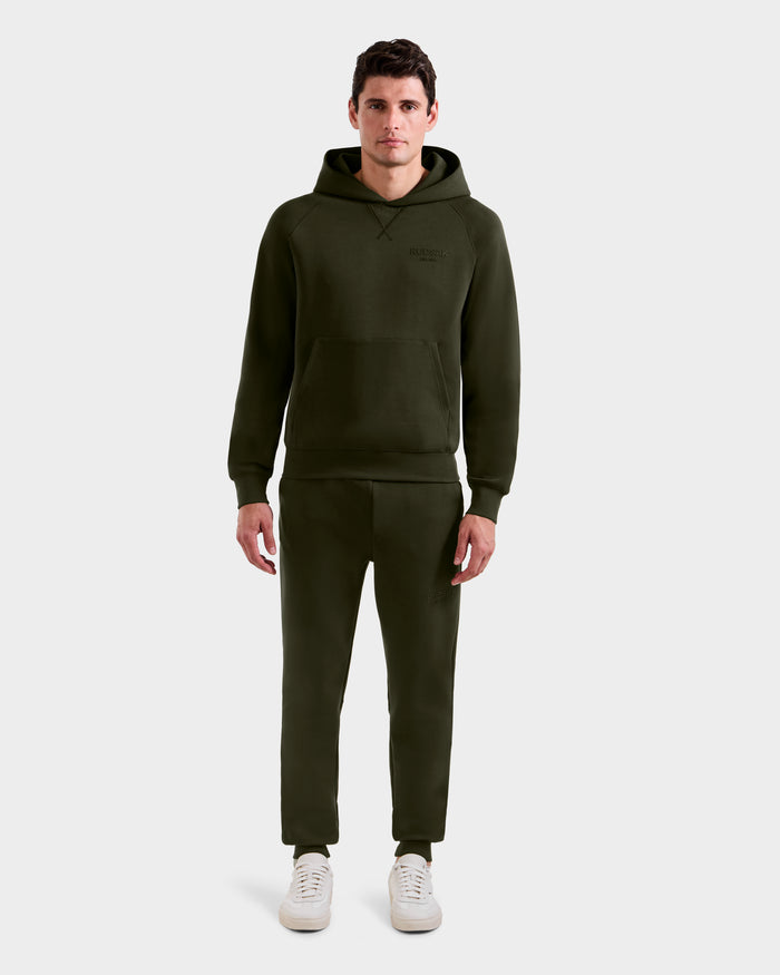 A man faces forward against a light gray background, wearing the Rudsak BARRY - 9524011 hoodie and matching jogger pants in Dark Olive (size M) with white sneakers.