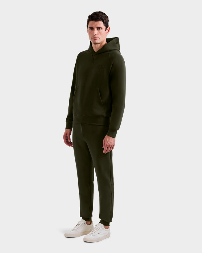 A man faces forward in a Rudsak BARRY - 9524011 unisex dark olive hoodie and jogger set, paired with white sneakers, against a plain light gray background.