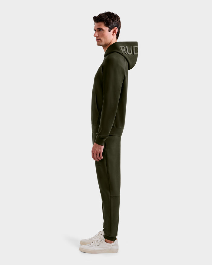 A man stands in profile against a light gray background, wearing the Rudsak BARRY - 9524011 hoodie in Dark Olive (size M), paired with matching joggers and white sneakers. The "RUD" logo is partially visible on the hood.
