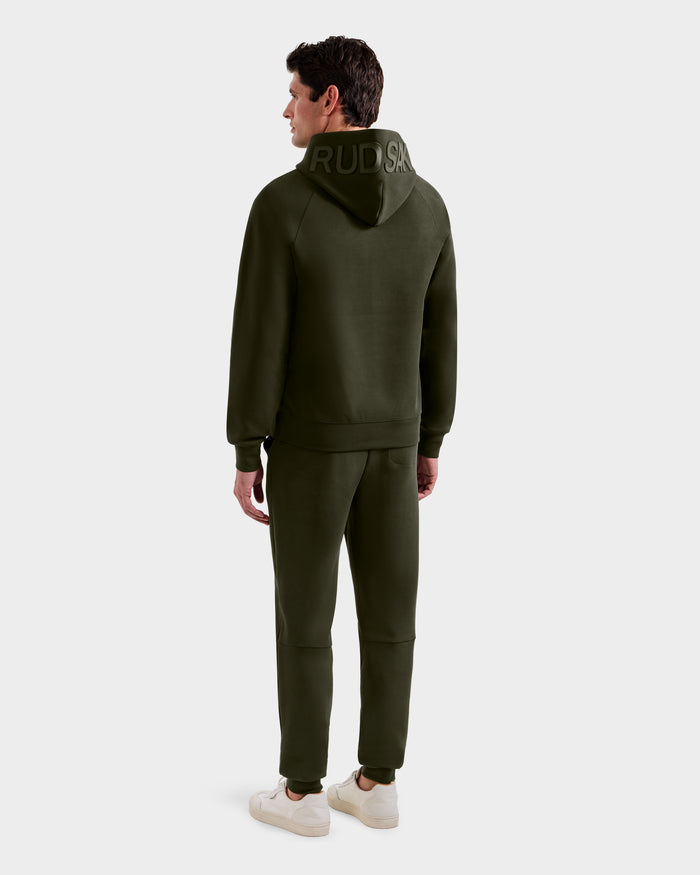 A man faces away, wearing the Rudsak BARRY - 9524011 hoodie in Dark Olive (M), paired with matching joggers and white sneakers. "RUDSAK" appears on the back of the hood, all set against a plain light gray background.